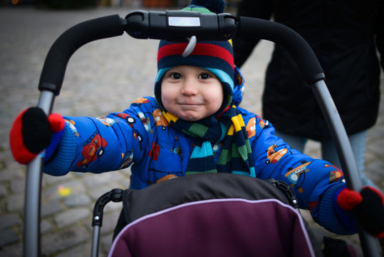 Cute Little Boy Rolls Baby Stroller On The Street. Beautiful Child Smiling And Acting Like An Adult. Portrait Of Active Kid Playing Outdoor