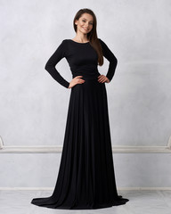 Young woman with long brunette hair dressed in elegant evening black maxi dress with long sleeves posing in studio. Gorgeous female model standing against white wall decorated with classic moldings.