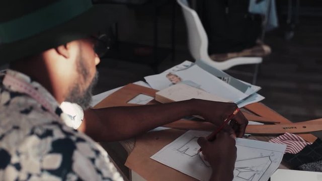 Over shoulder shot of a young, stylish, Afro-American designer sketching out a new fashion item with pencil in a cozy, modern office. Fashion, style, luxury. Male portrait
