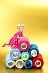 The girl sits at the top of a huge mountain of coils with sewing threads for 3 years. The little beautiful blonde girl climbed onto the pyramid of bright colored threads.