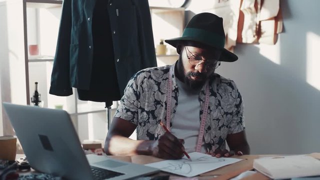 Male fashion designer draws, colors sketches for his future fashion collection. Modern art, creative process. Cozy office, mannequin, crammed table. Male portrait