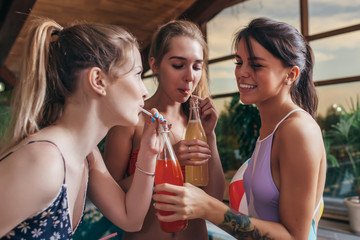 Three happy girlfriends in swimwear drinking juice from bottles at swimming pool in spa center