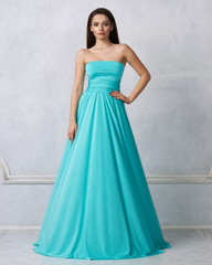 Fototapeta na wymiar Beautiful long haired young woman dressed in stylish turquoise blue bandeau maxi dress posing against white wall on background. Elegant brunette female model demonstrating evening outfit in studio.