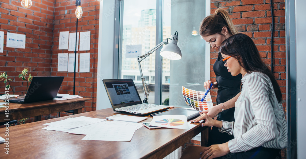 Wall mural young women working on a new web design using color swatches and sketches sitting at desk in modern 