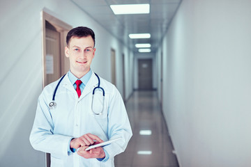 doctor in white medical coat  is using a tablet and smiling while standing against window