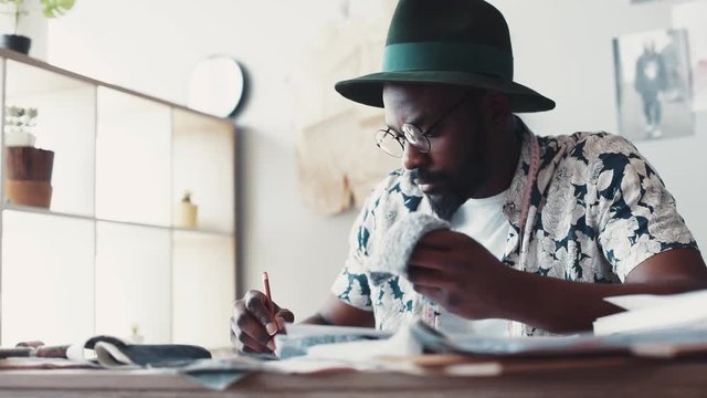 Trendy Afro-American man probes different kinds of fabric, decides what exactly to choose for the design he is sketching. Talented artist, modern culture, in vogue. Slow motion, close up view, male
