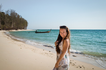 Fototapeta na wymiar Young woman in dress is walking on the beach on the coast of the ocean. Smiling girl is enjoying sunny summer day and looking in camera. Tropical tavel destination