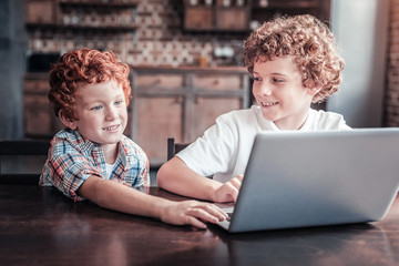 Modern generation. Positive cheerful nice boy sitting at the laptop and looking at his brother while spending time with him