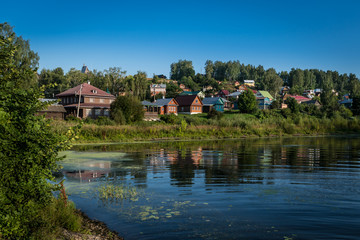 Fototapeta na wymiar Scenic landscape with traditional buildings in Russian village on the riverbank. Background of nature with green trees, grass and hills. Clear sky
