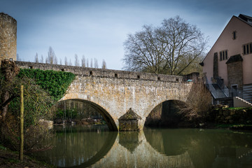 Beautiful antique bridge with plants on top over the river at old town. Reflections of constructions on the water. Authentic architecture in lower city in Luxembourg