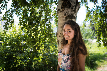 Fototapeta premium Young attractive woman stands near the tree in the park on the background of green leaves and sun rays. Beautiful cheerful girl is smiling and standing under the birch in summer warm day