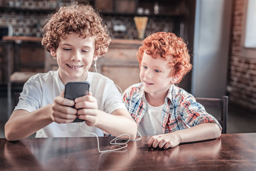 Music playlist. Happy positive nice boy sitting at the table and looking at the smartphone screen while choosing the song