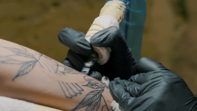 tattooer in violet clothing making tattoo on the man's arm of her client with gun in studio. Young business woman in workshop. 4k Close up view