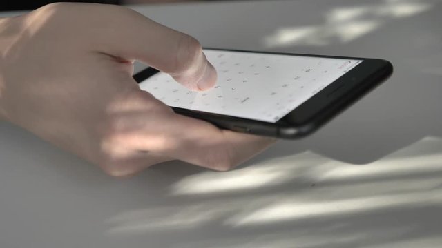 Men's hand scrolling calendar on a big black smartphone, typing, sunlight on the screen, reflection, concept. A sunny table in the background. 60 fps