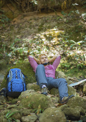 Young tired hiker with backpack sitting on the rock in forest while resting after active walk. Travel concept