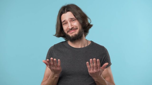 Portrait of handsome smiling guy with beard and brown hair embarrassing and waving hand in hesitation, over blue background. Concept of emotions