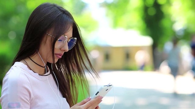 Attractive woman in glasses with smartphone and earphones listens to music. Girl with smartphone listens to music in park