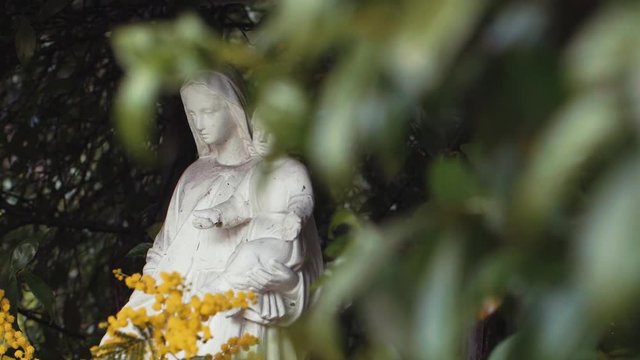 Panning shot revealing a small stone sculpture of the Virgin Mary holding Baby Jesus in a small green bush in a garden in Rome