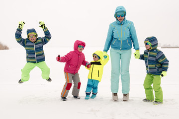 Happy young mother in blue ski suit wearing sunglasses with funny children in bright winter clothes jumping for joy on shore of icy lake. Wonderful winter vacation for whole family. Kids fun together