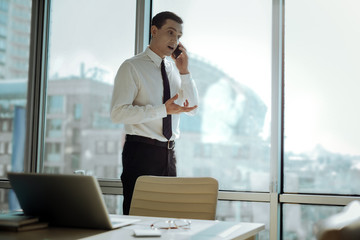 Important negotiations. Pleasant business man standing near the window in his office and talking on the phone emotionally, negotiating with his partners