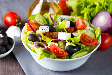 Fresh Greek salad made of cherry tomato, ruccola, arugula, feta, olives, cucumbers, onion and spices. Caesar salad in a white bowl on wooden background. Healthy organic diet food concept.
