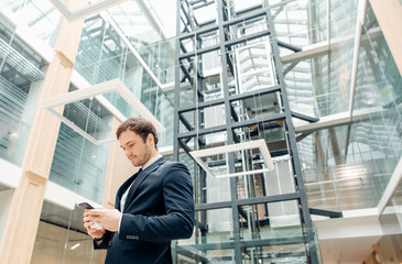 Fototapeta premium Coffee break on go. confident man holding coffee cup and using his smart phone while walking in hall