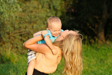 Young blonde mother in playing with her child in the green lawn. Happy fun summer photo. Smiling laughing boy with his mom. Sunny day outdoors in nature. Warm hug and embrace.
