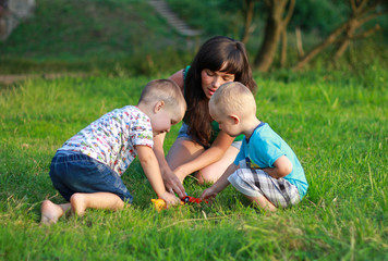 Naklejka na ściany i meble Mother with dark hair is playing with two children in the green lawn. Happy fun summer photo. Smiling laughing boys with his mom. Sunny day outdoors in nature. Kids and mom are hugging.