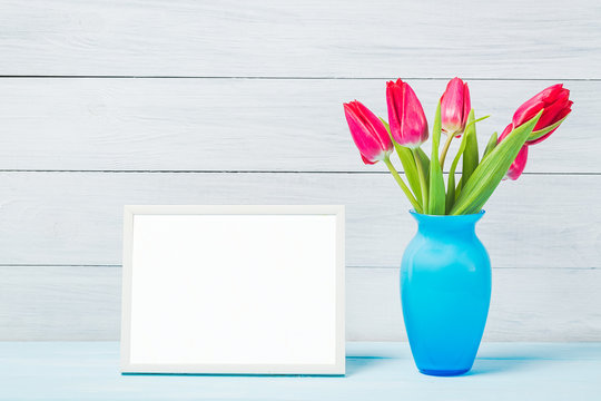 Colorful red spring tulip flowers in nice blue vase and blank photo frame on light wooden background as greeting card. Mothersday or spring concept