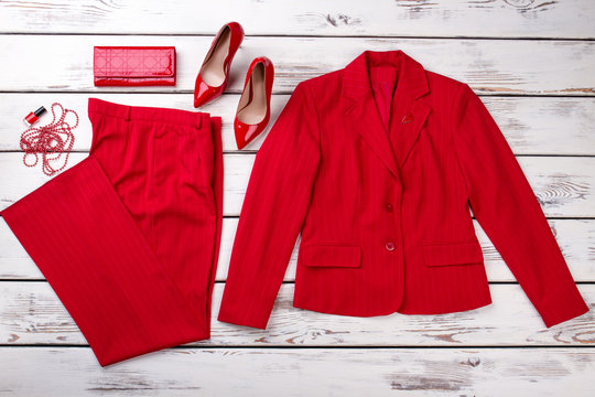 Flat lay red women suit and accessories. Jacket, trousers, wallet, shoes, necklace beads and lacquer. Bright wooden desks surface background.