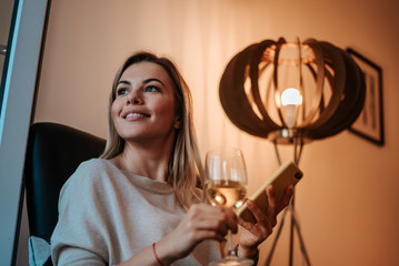 Close-up image of smiling woman enjoying glass of white wine at evening. Using smartphone.