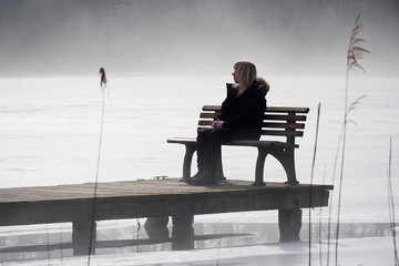 Frau sitzt alleine auf einer Bank am See im Winter mit Nebel