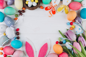 Fototapeta na wymiar Easter composition with colored eggs, rabbit ears, candy, nest and tulips on a white wooden background.