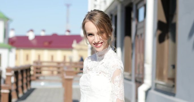 Elegant girl with hairdo in white lace design dress walking smiling in street sunny spring day back. Young woman wearing festive clothing looks camera make-up skin care. Female beauty happiness

