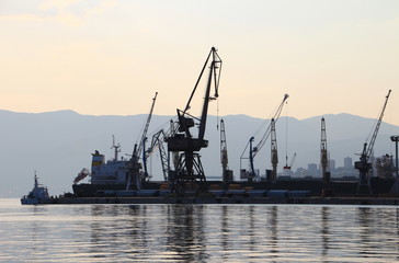 Obraz premium Silhouettes of port cranes at quayside and large bulk carrier ship in Croatian harbour at sunset in evening light with Istrian Ucka mountains, Rijeka city Kvarner Gulf Croatia Balkan Peninsula Europe