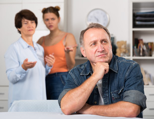 Woman and her daughter are sympathying their sad father who is sitting at the table