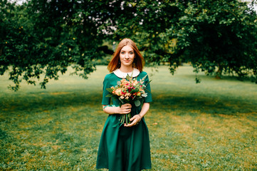 Young happy red haired girl with bouquet of summer flowers enjoying inner peace in green dress at nature. Little child playing in wonderland park. Pretty woman portrait outdoor. Lovely smiling face.