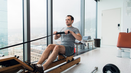 Rowing with power. Side view of young man in sportswear doing rowing in front of window at gym.