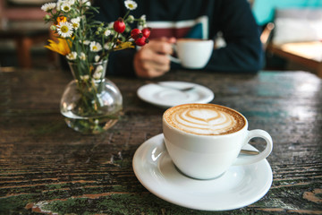 Two cups of coffee and a vase of flowers on a wooden table, the man holds in her hand one cup of coffee in the background. A photo indicates a meeting of people and a joint pastime.
