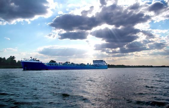 The rays of the sun make their way through the cumulus clouds in the blue sky and illuminate the navigable river along which the tanker sails