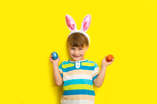  Cute positive little boy wearing bunny ears holding colorful Easter eggs on a yellow background.
