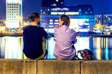 Young couple watching a sunset behind a cityscape sitting on a quay of a river.