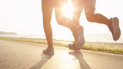 LOW ANGLE, SUN FLARE: Young athletic woman and man run along sunny ocean road.