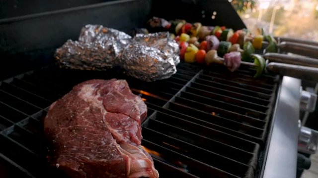 Large beef steak grilling on a bbq with a man turning vegetable kebabs