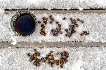 Mug of black espresso coffee stands in the snow, a wooden bench in the park, winter, scattered beans