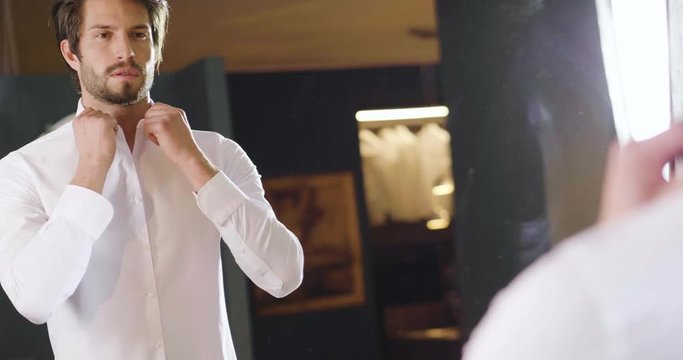 A handsome man in his loft dresses his sexy white shirt in a sexy way, looks into his room and smiles with a sure look at himself. Concept of: elegance, business, work.
