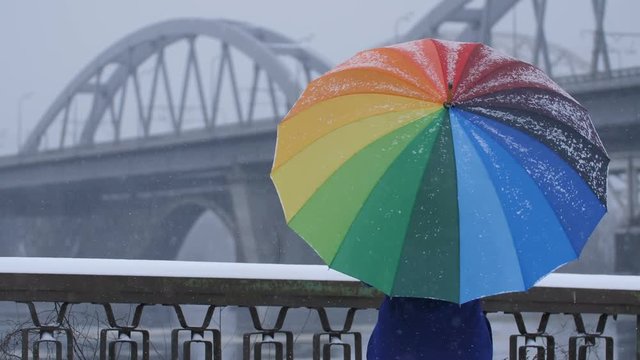 Lonely female spinning colors rainbow umbrella in winter during snowfall and. Urban bridge architecture at the background on a gloomy winter day. Hope, faith and dreams concept.