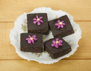 Four homemade brownie squares with chocolate frosting and pink and yellow piped flowers on a white flowered plate against a wood table.