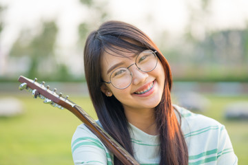 Asian teen girls  with a guitar at the shoulder in the lawn. She is wearing braces  and wear eyeglasses.