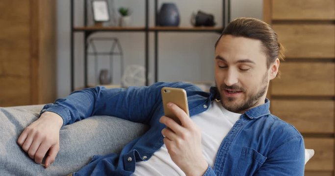Portrait of the attractive man in jeans shirt resting on the sofa and texting and taping on the smartphone in the living room. Indoors